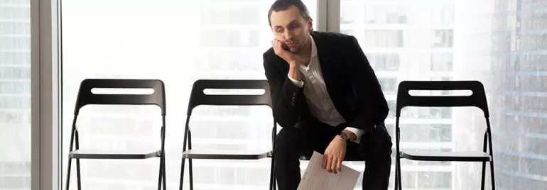 Man in a suit sitting on a chair, resting his head on his hand, looking tired, with empty chairs beside him.