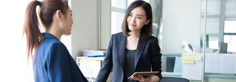 Two women in business attire having a conversation in an office.