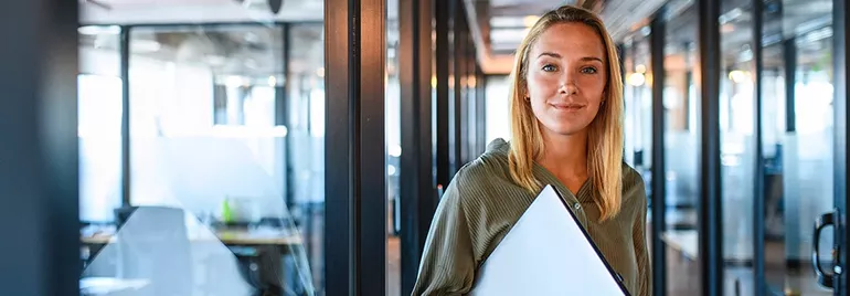 Woman standing in a modern office corridor holding a laptop.
