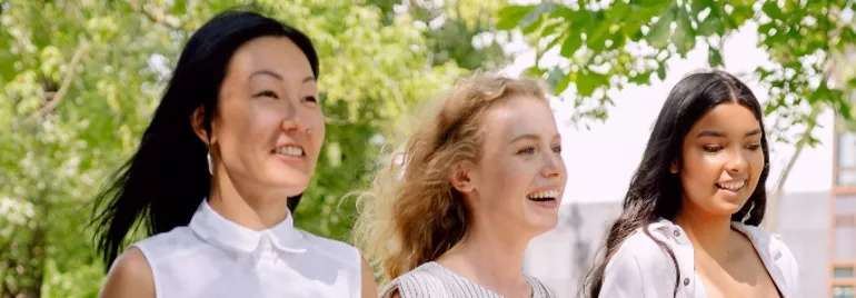 Three women smiling and walking outdoors under leafy trees.