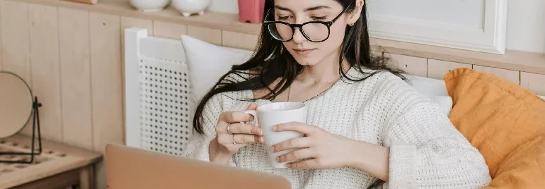 Woman with glasses sipping coffee while using a laptop.