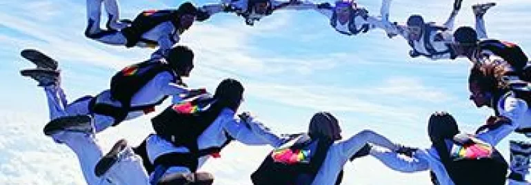 Group of skydivers forming a circle in the sky against a backdrop of clouds on a sunny day.