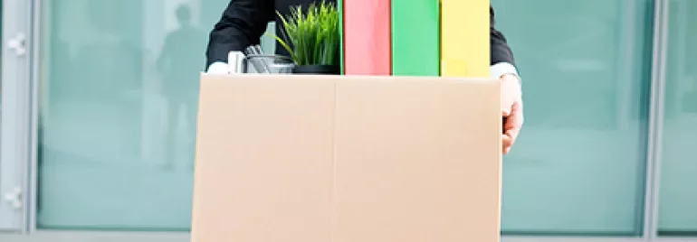 Person holding a cardboard box with colorful binders and a potted plant.