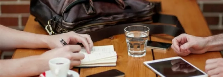 Two people at a table with coffee, notebook, tablet, glass of water, and a bag.