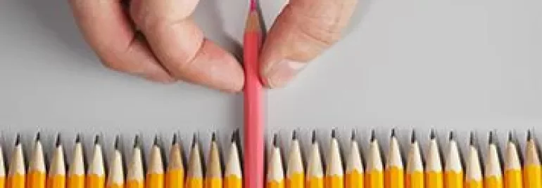 A hand picking a pink pencil out of a row of yellow pencils.