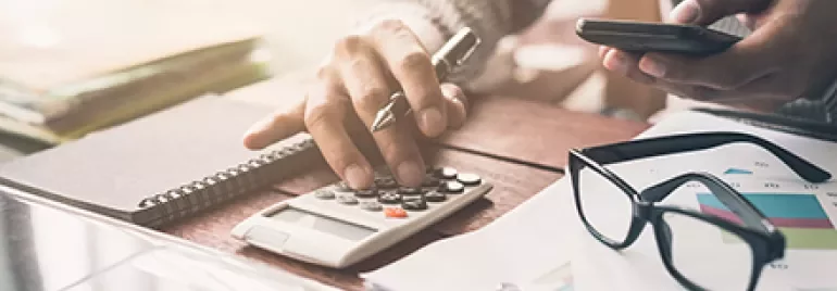 Person using a calculator at a desk with a smartphone, notepad, and glasses.
