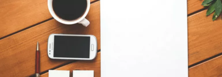 Workspace with coffee, smartphone, pen, and blank paper on a wooden table.