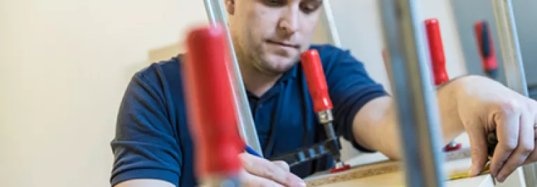 Man measuring and marking a wooden board using a pencil and clamps.