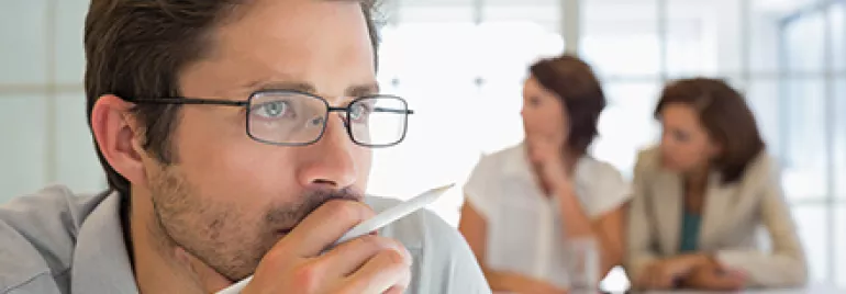 Man thinking with pen in hand, two women in background.