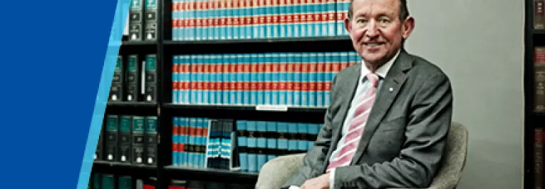 A man in a suit sits in a chair next to shelves filled with books.