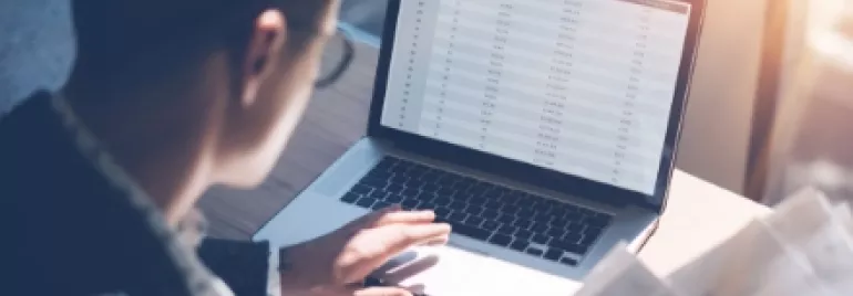 Person analyzing data on a laptop with spreadsheets and documents in hand.