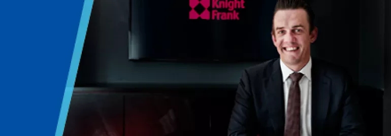 Businessman in suit smiling at desk with Knight Frank logo on screen behind.