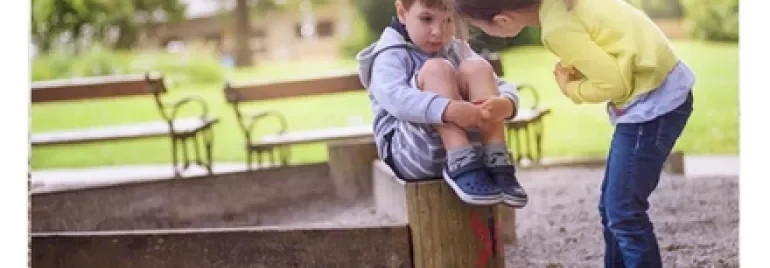 Child sitting on a wooden post while another child leans in, in a park setting.
