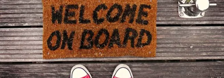 Doormat on wooden dock reads 'Welcome On Board' with red sneakers visible.