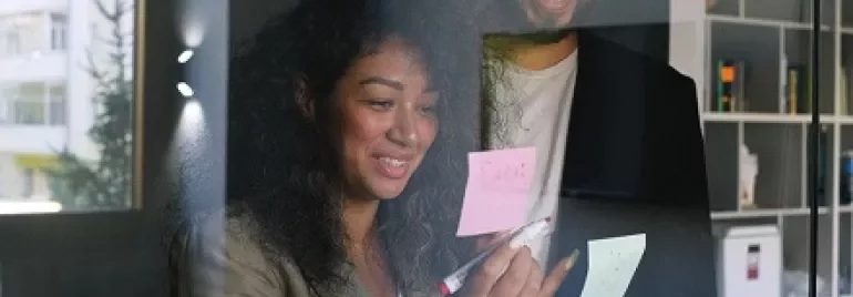 Two colleagues smiling while brainstorming with sticky notes on glass.