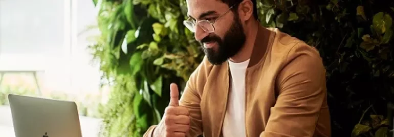 Man giving thumbs up while using laptop and phone in a cafe.