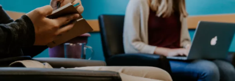 Person taking notes while another works on a laptop in a waiting room.