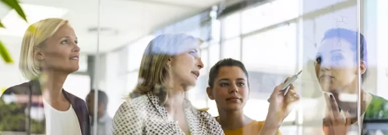 Business meeting with four women discussing in front of a glass board.