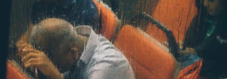 Three people sitting on a bus in the rain, viewed through a wet window.