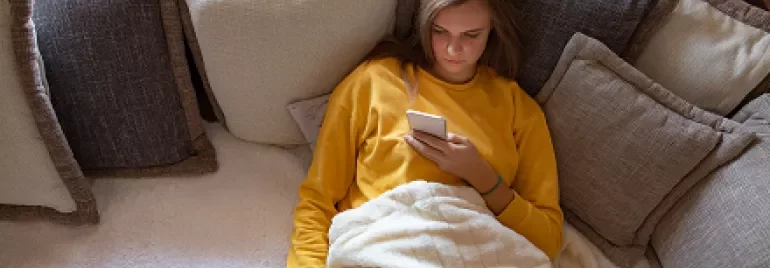 Person in yellow shirt lying on a sofa with pillows, looking at a phone.
