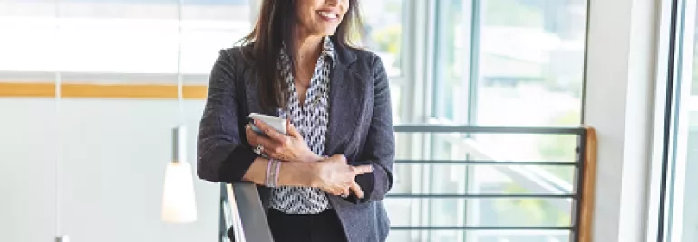 Woman standing in a modern office, holding a phone and smiling.