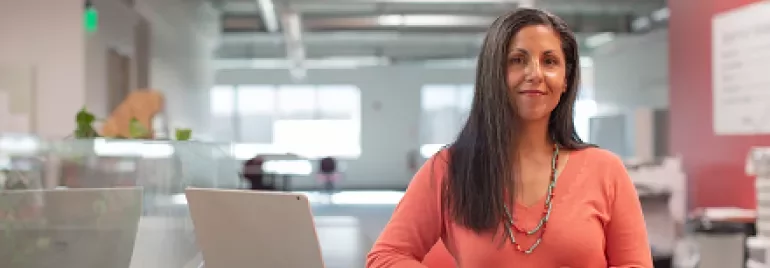 Woman in orange sweater standing by a laptop in a modern office.