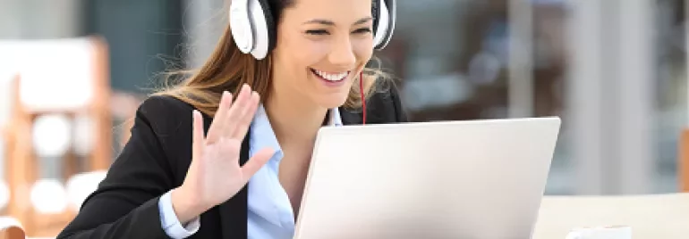 Smiling woman with headphones waves during a video call on a laptop.