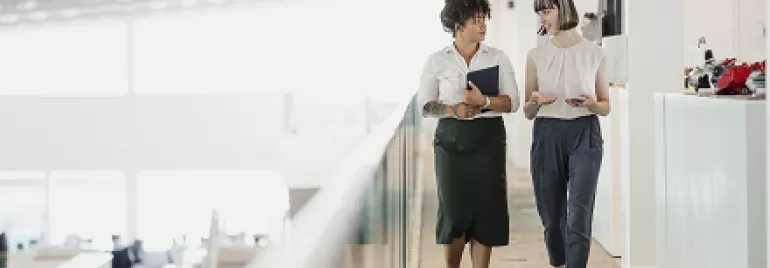 Two women walking and talking in a modern office hallway.