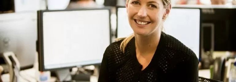 Smiling woman at office desk with computer monitors in the background.