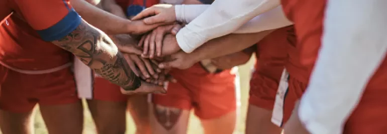Rugby team huddles with hands together before a match, wearing red uniforms.
