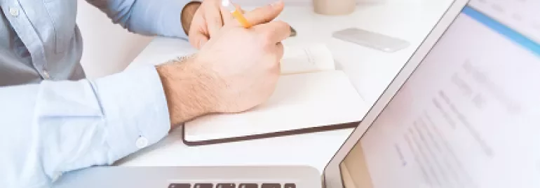 Person writing in a notebook at a desk with a laptop and plant.