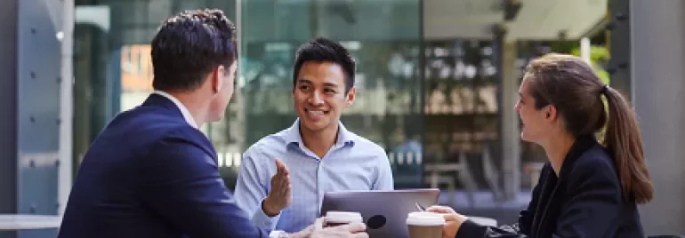 Three people having a meeting at an outdoor cafÃ© table, smiling with coffee cups and a laptop.