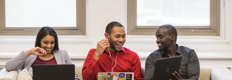 Three people sitting on a couch using laptops, smiling and talking to each other.