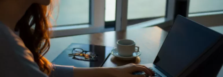 Woman typing on a laptop with a cup of coffee on a wooden table.