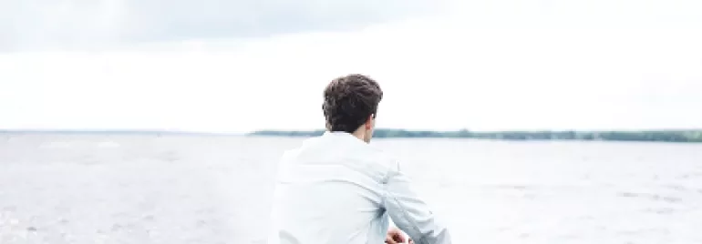 Person sitting by a beach looking at the water under a cloudy sky.