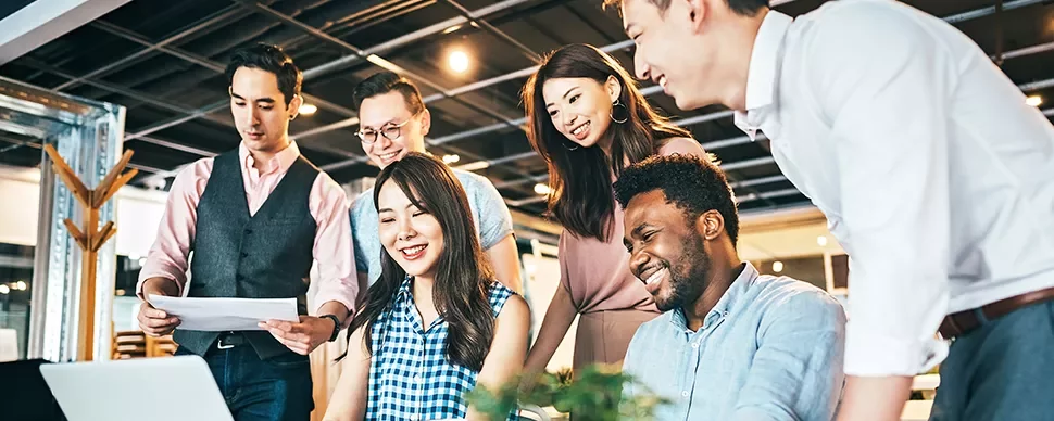 A group of colleagues collaborating at a desk, smiling and looking at a laptop.