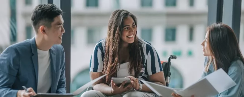Three people in a meeting, smiling and discussing with papers and tablets.