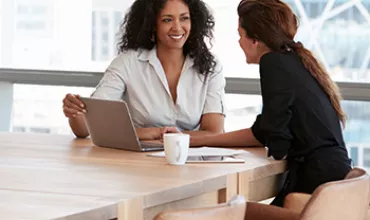 Two women discussing work at a table with a laptop and coffee mug.