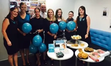 Group of people in an office with blue balloons and snacks on tables.