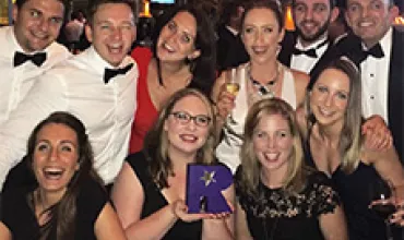 Group of smiling people in formal attire at an event, holding a purple award.