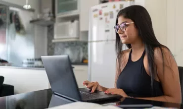 A woman with glasses working on her laptop in a kitchen.
