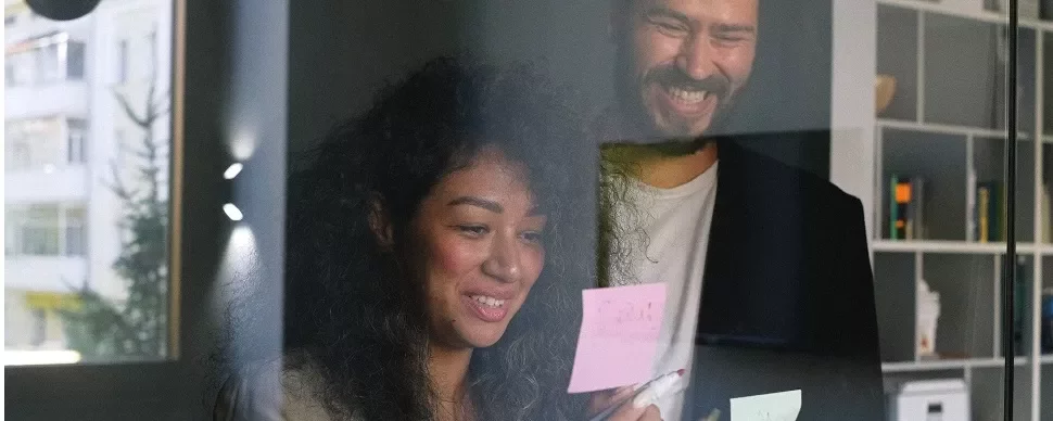 Two people smiling and reading sticky notes on a glass wall in an office.