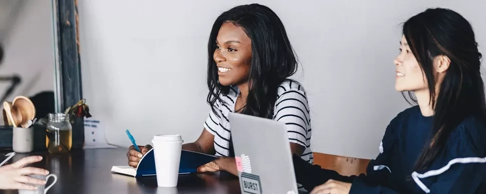 Two women at a table with laptops and coffee, engaged in discussion.