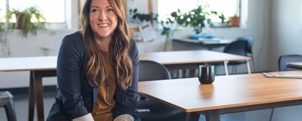 Smiling person sitting in a modern office with wooden tables and plants.