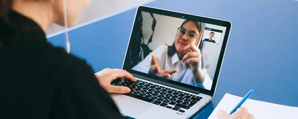 Person on laptop video call taking notes with a pen.