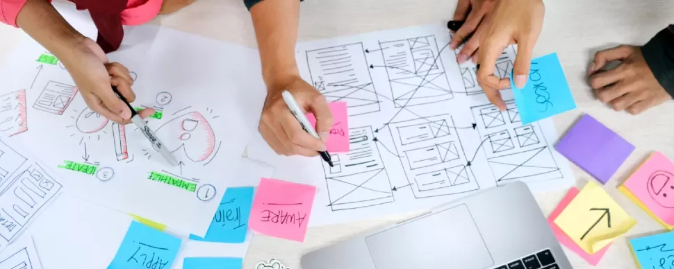 People brainstorming at a desk with wireframe sketches and colorful sticky notes.