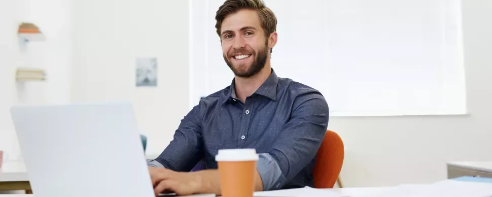 Smiling man in a blue shirt working on a laptop at a desk with a coffee cup.