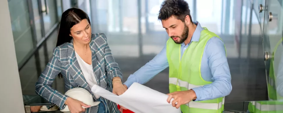 Two professionals reviewing building plans in an office corridor.