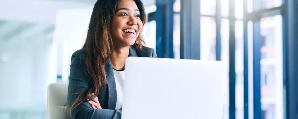 Woman in a suit smiling while working on a laptop in a bright office.