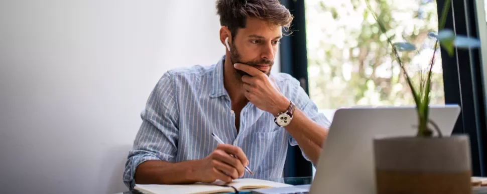 Man wearing earphones working on a laptop while taking notes.
